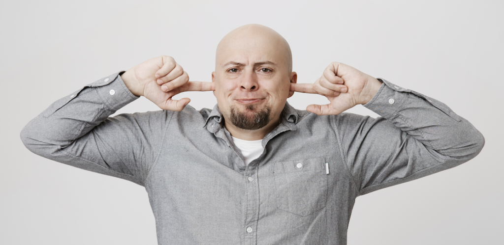 A bald man with a beard is seen pressing his index fingers into his ears with a frustrated expression on his face. He is wearing a gray button-up shirt and standing against a plain white background. The gesture suggests he is trying to block out noise or sound, potentially indicating discomfort or irritation.